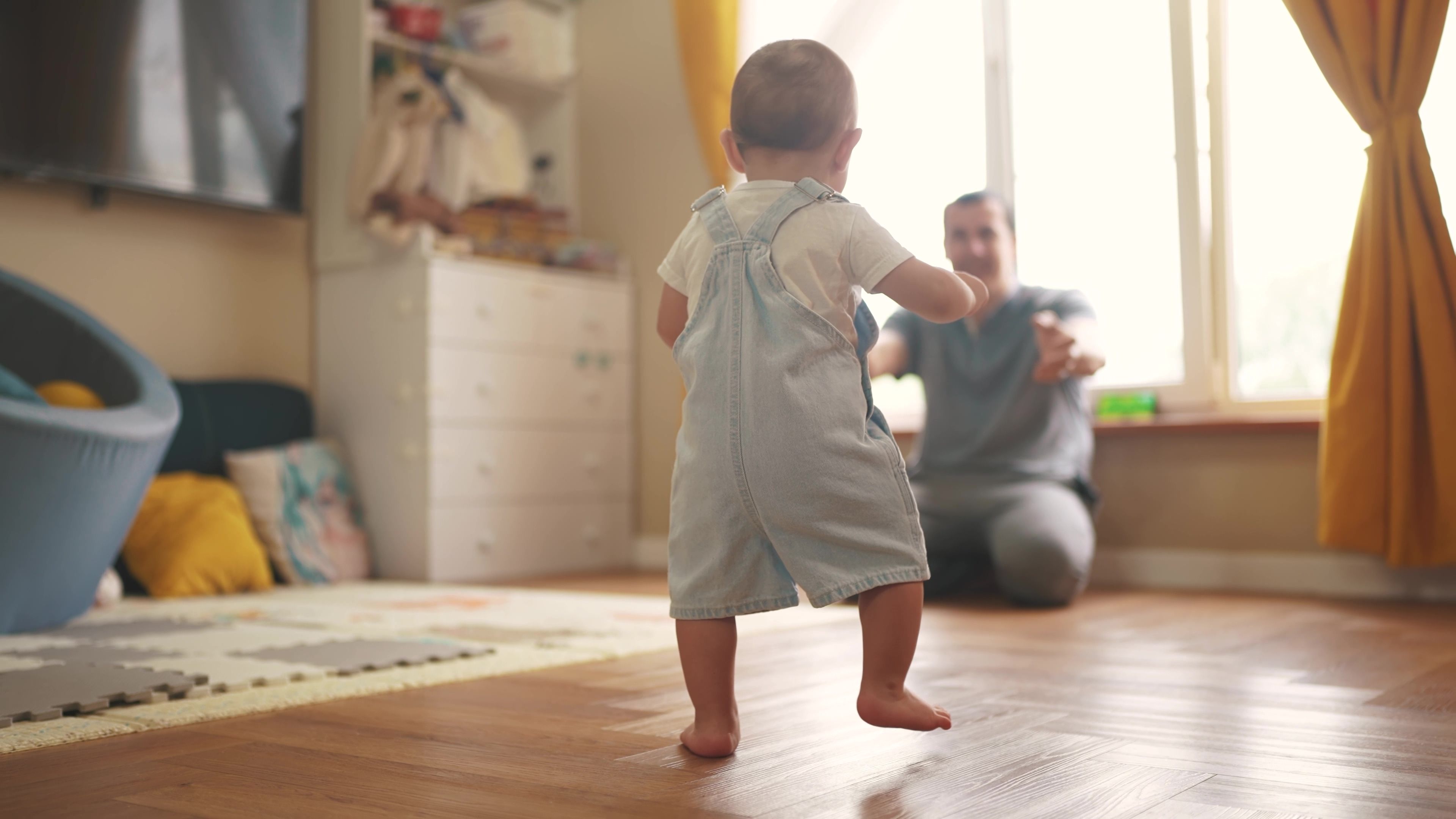 Toddler taking first steps in a sunlit nursery with parent encouraging from behind